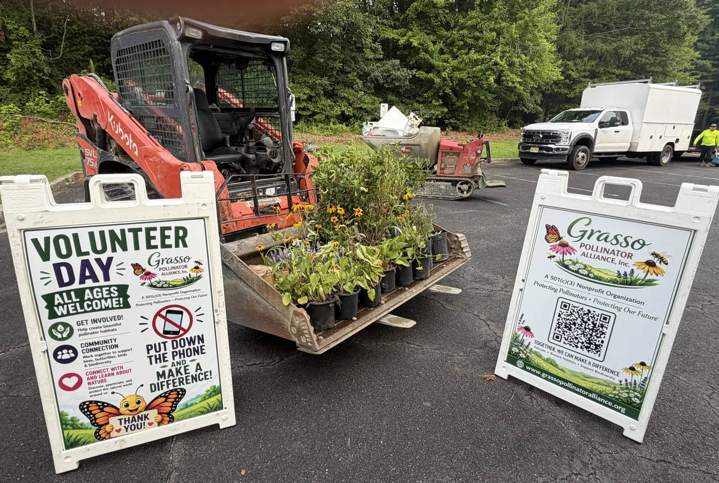A Volunteer Day A-frame sign and a Grasso Pollinator Alliance QR sign beside a track loader and a pallet of native plants